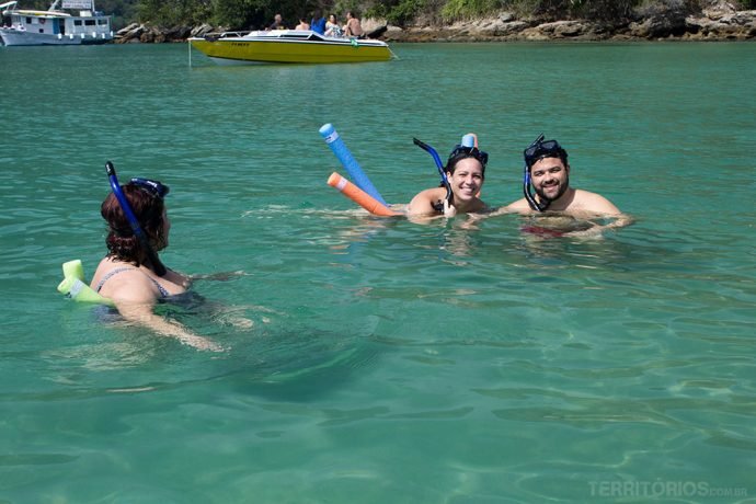 Três pessoas aproveitam o mar parecendo lagoa com snorkel. Ao fundo lancha para descobrir Ilha Grande
