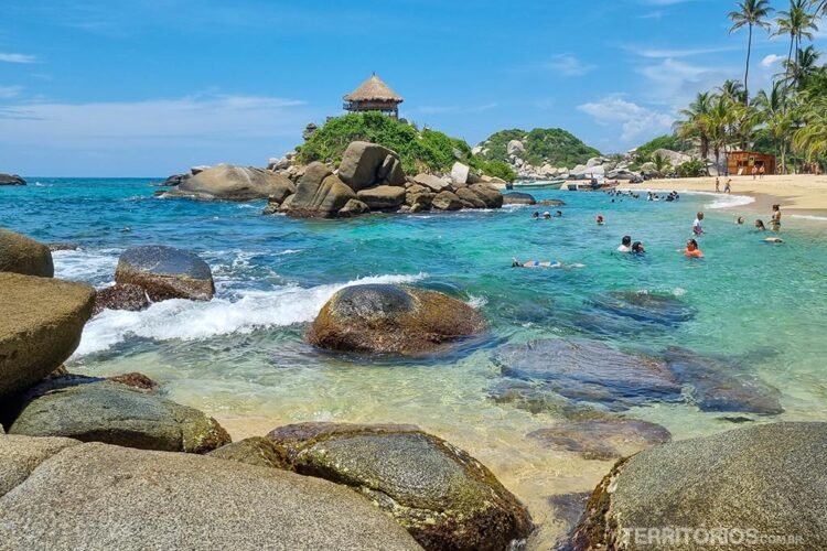 Praia com pedras e coqueiros no mar verde e transparente do Caribe Colombiano. Pessoas na água e um quiosque no topo da rocha