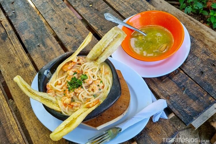 Prato servido com massa, camarão e banana chips e outro com sopa em mesa de madeira