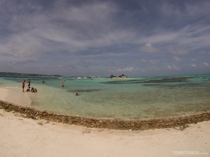 Céu azul com nuvens, mar verde com corais escuros e pessoas aproveitando o dia na praia. Barreira de pedras para contar a água. 
Vista de Haynes Cay para o Acuario durante o passeio de lancha para conhecer San Andrés.