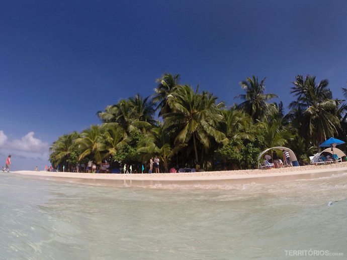 Ilha com coqueiros e barracas com turistas, céus azul e poucas nuvens ao fundo. 
Johnny Cay visto de dentro d’água durante o passeio de lancha para conhecer San Andrés. 