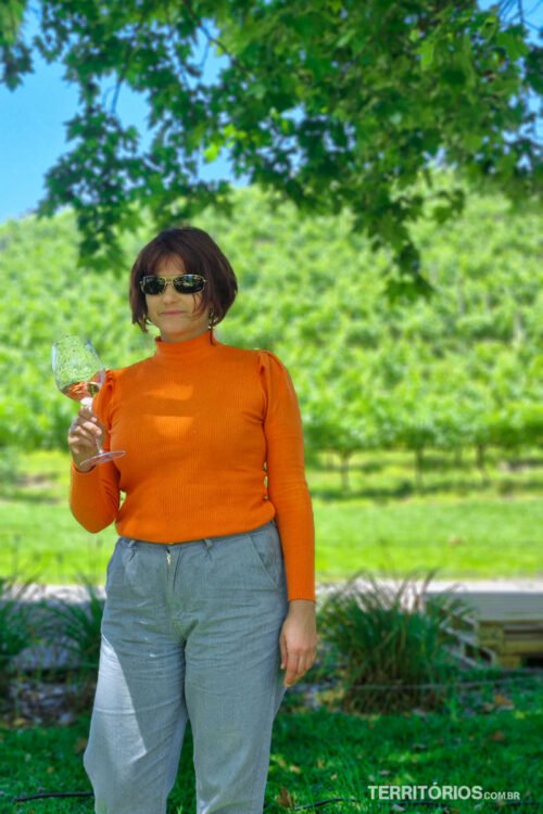 Mulher veste blusa laranja e calça jeans, leva taça de vinho branco na mão e fundo ver na natureza do Vale dos Vinhedos