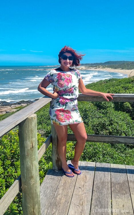 Mulher morena posa com vestido curto branco floral e óculos de sol. Cabelos ao vento. Ao fundo vegetação e praia. Céu azul.