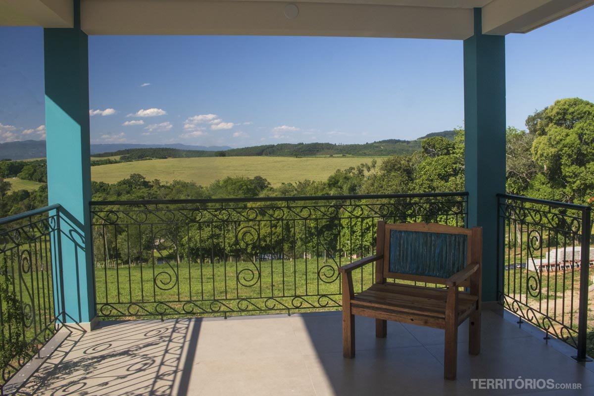 Um cadeira na sombra. Varanda com vista para campo verde em dia de céu azul é opção de hospedagem no Vale Germânico