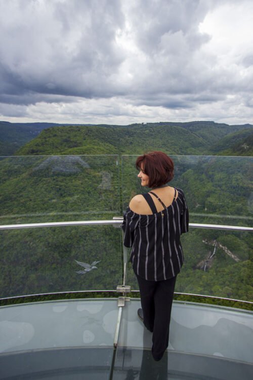 Mulher vestida de preto, de costas, sorri olhando a paisagem da plataforma de vidro