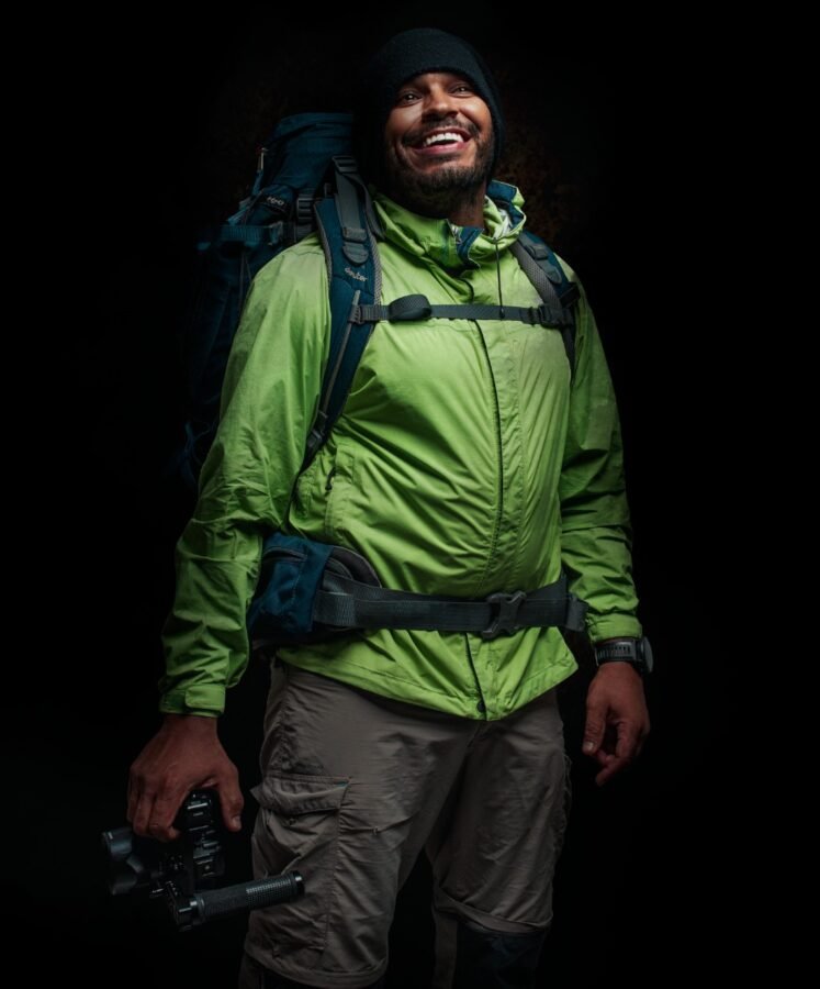 Homem veste agasalho verde, touca escura, calça beje e mochila pronto para trilhas, na mão leva uma câmera fotográfica. Drone da Montanha