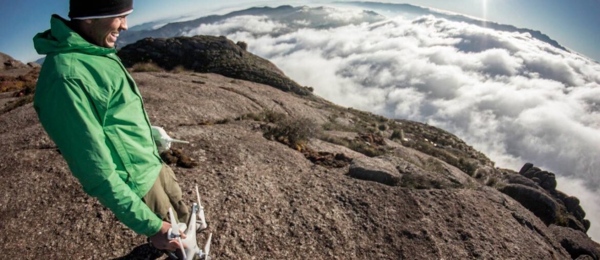 Homem com drone da mão sorri e observa a paisagem acima das nuvens do alto de uma montanha