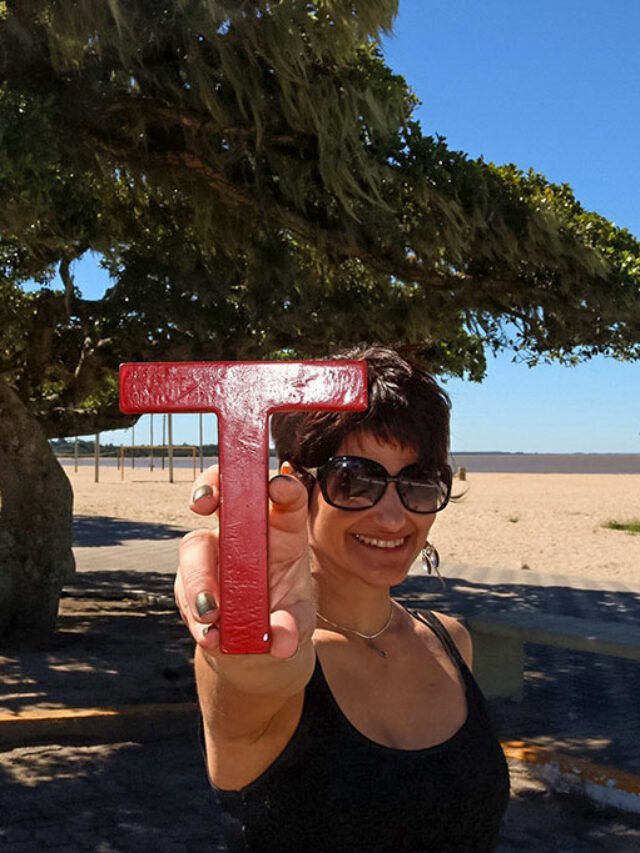 Mulher segura letra T vermelha, ao fundo figueira e praia com areia