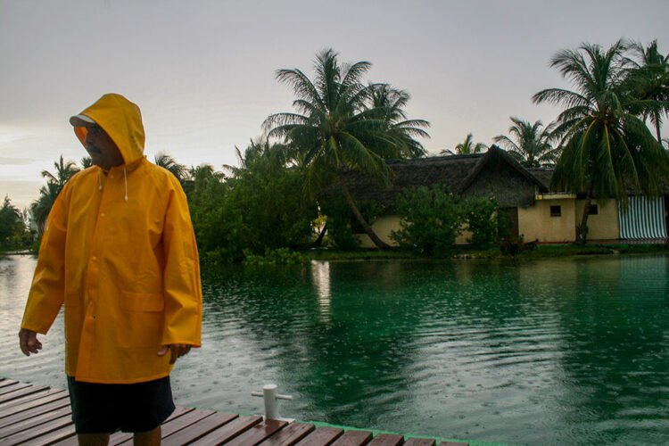Dia de chuva no Caribe, homem veste capa amarela, ao fundo casa e coqueiros, mar verde