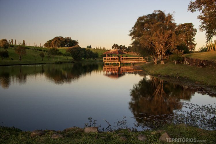 Lago com reflexo rodeado por gramado e árvores no final de tarde. Gazebo no lago