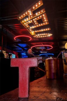 Letra T (Tesão) de madeira em mesa de bar com neon no teto escrito Hello