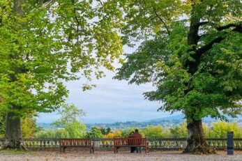 Duas pessoas sentadas em um banco apreciam a vista para as montanhas entre árvores do Pays de Gex