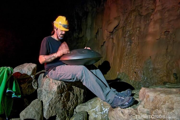 Homem veste capacete e toca instrumento musical de metal no interior de uma caverna