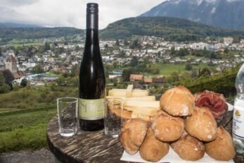 Mesa com pães, queijos, copo de vidro e garrafa de vinho. Ao fundo a cidade de Spiez