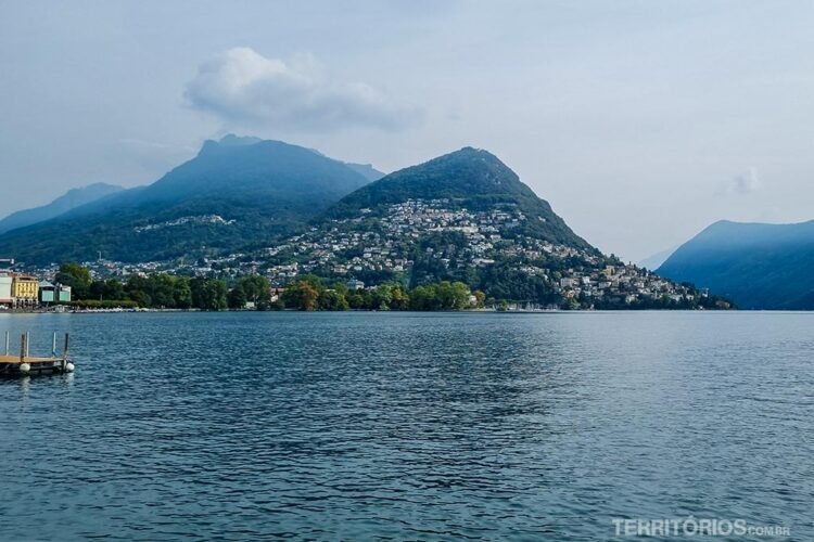 Céu entre nuvens, montanhas com casas nas encostas e lago Lugano