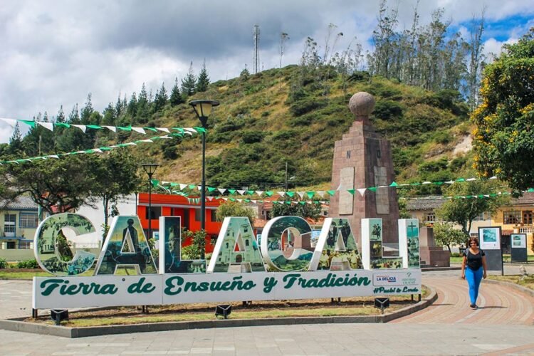 Praça de Calacalí e o monumento equatorial. Mulher caminha pelo calçamento de pedra com calça azul e blusa preta. Colina verde ao fundo e céu nublado