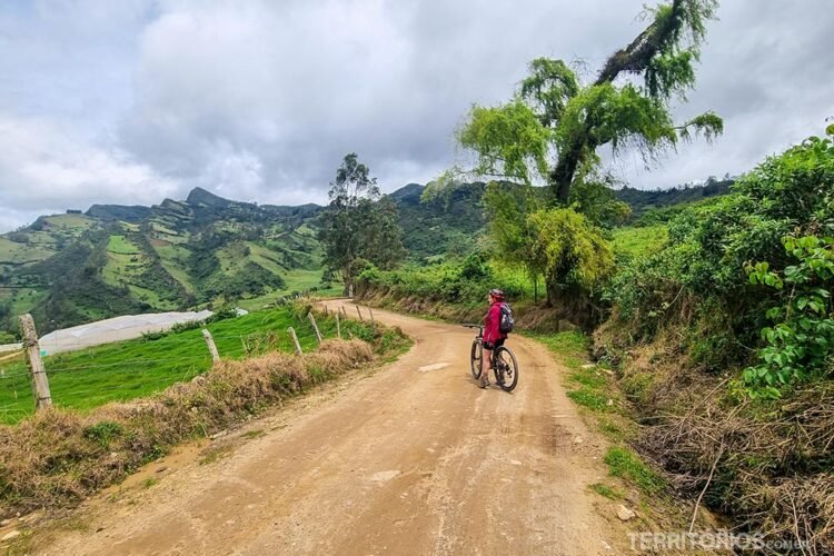 Pessoa com casaco vermelho de bicicleta em estrada de terra, dia nublado, vegetação