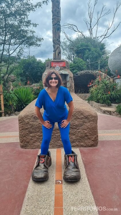 Mulher veste conjunto azul em frente ao monumento da Linha do Equador, posa colocando um pé em cada sapato demarcando o hemisfério. Céu nublado, monumento de pedra e vegetação atrás