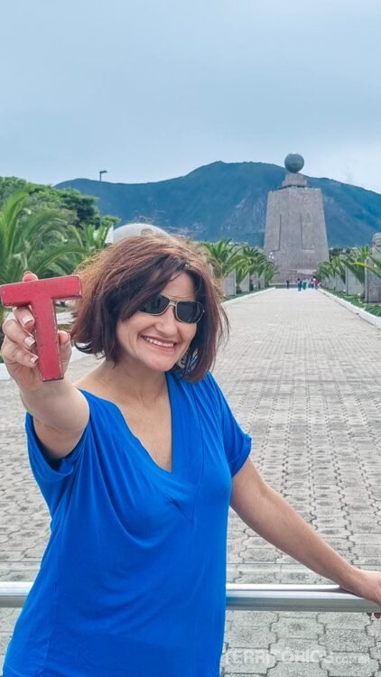 Mulher veste azul e óculos, segura uma letra T vermelha na mão. Ao fundo Monumento da Linha do Equador e céu nublado.