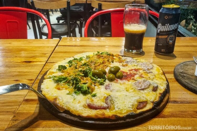 Pizza servida em mesa de madeira, copo e lata de cerveja preta. Dicas de onde comer em Buenos Aires