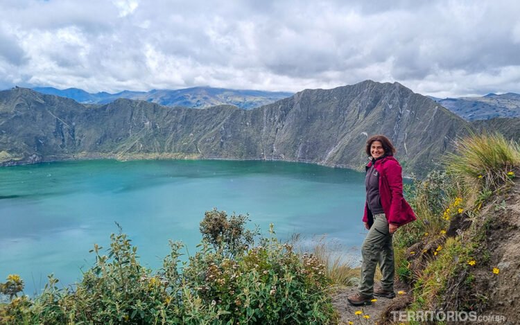 Mulher veste calça bege e casaco magenta sorrindo na borda da cratera do vulcão Quilotoa