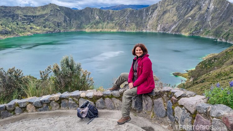 Mulher veste calça bege e casaco magenta sorrindo no muro em frente ao lago do vulcão Quilotoa