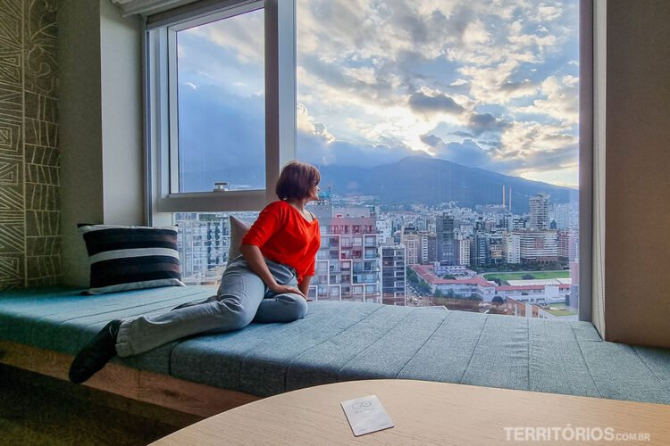 Mulher veste blusa laranja e calça jeans, se debruça na janela com vista para a cidade sentada em sofá azul.