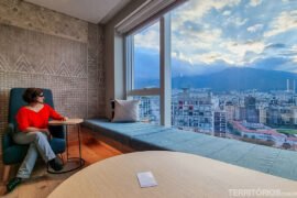 Mulher veste blusa laranja e calça jeans, sentada em poltrona azul olhando a janela com vista para a cidade.