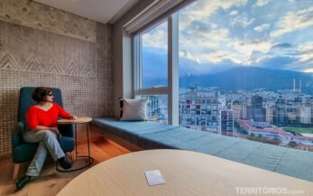 Mulher veste blusa laranja e calça jeans, sentada em poltrona azul olhando a janela com vista para a cidade.