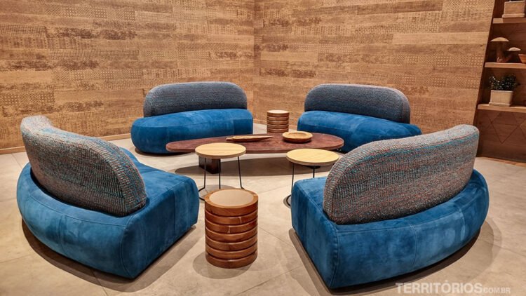 Quatro sofás azuis formam sala de estar em círculo com mesas de centro e de apoio. Ambiente rústico com paredes de madeira texturizada e estante.