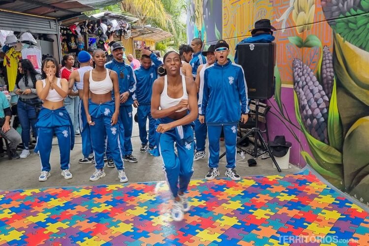 Homens e mulheres usando uniforme azul dançam na rua. Grafite na parede, tapete colorido no chão.