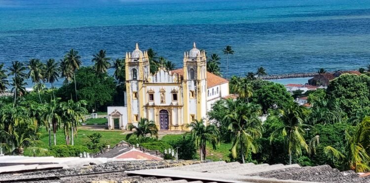 Igreja colonial rodeada por coqueiros e mar azul ao fundo