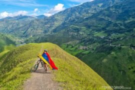 Turismo na Colômbia: dicas aos viajantes Mulher em bicicleta envolta em bandeira em cenário verde de montanhas para promover o turismo na Colômbia