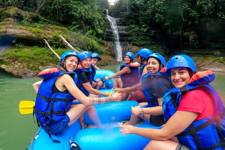 Grupo faz pose durante rafting no rio Guejar. Todos vestem colete salva-vidas e capacetes azul, carregam remos e estão em bote inflável azul. Ao fundo cachoeira e densa vegetação verde.