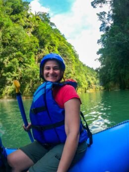Mulher sorri durante rafting no rio Guejar. Veste colete salva-vidas e capacetes azul, blusa rosa e carrega remo. Sentada em bote inflável azul. Ao fundo densa vegetação e rio verde.