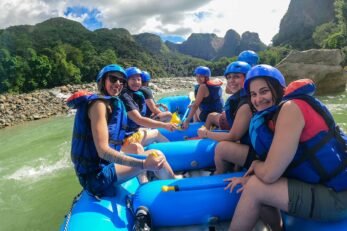 Pessoas em bote inflável vestindo equipamentos de segurança navegam por rio em meio a natureza