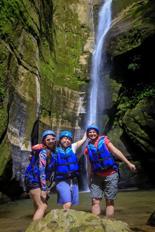 3 mulheres vestem colete salva-vidas e capacetes azuis. Ao fundo cachoeira e paredão de rocha e vegetação verde