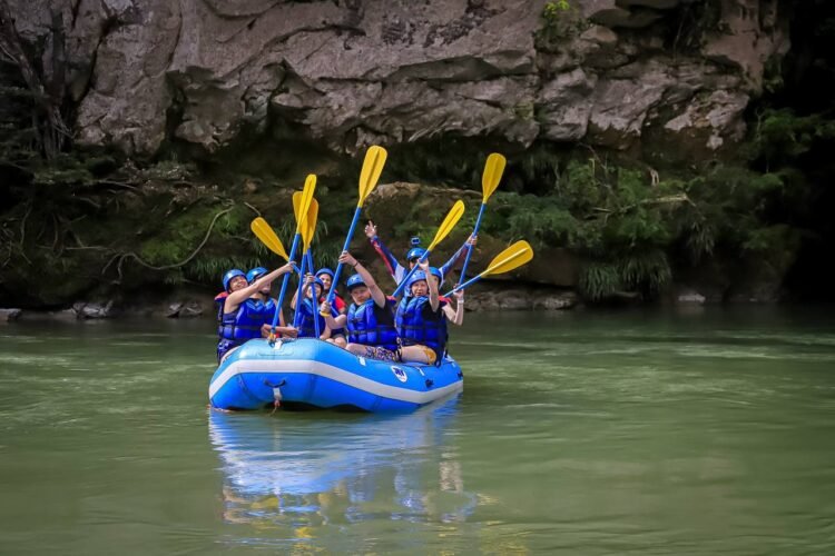 Grupo faz pose durante rafting no rio Guejar. Todos vestem colete salva-vidas e capacetes azul, carregam remos e estão em bote inflável azul. Ao fundo rocha e densa vegetação verde.
