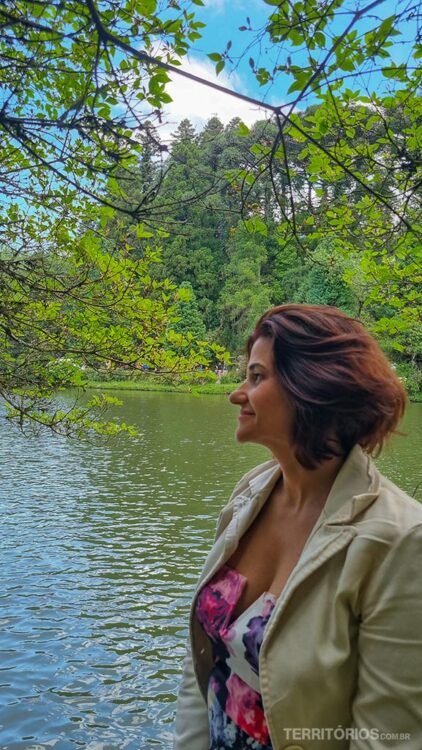 Mulher de cabelo castanho com blaser bege e blusa florida em frente ao lago, entre as atrações em Gramado