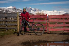 Bike elétrica te leva mais longe com menos esforço Mulher veste vermelho e segura bicicleta em frente a uma porteira bordô. Montanhas nevadas ao fundo