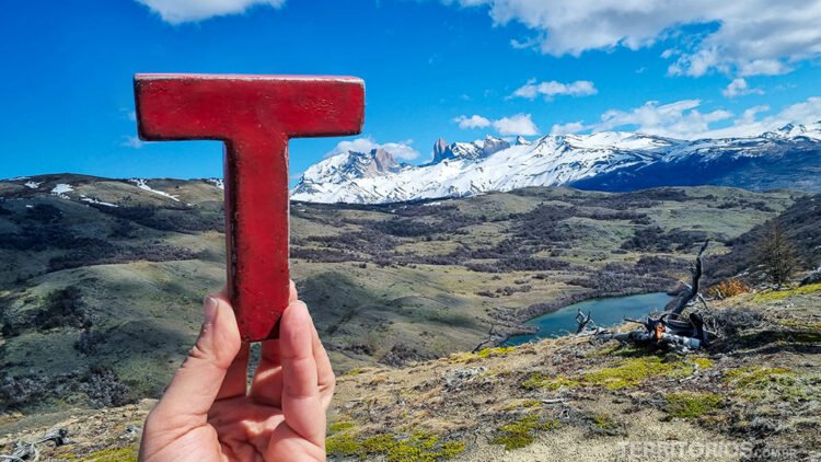 Mão segura letra vermelha na paisagem com montanhas nevadas, campos e lago verdes neve