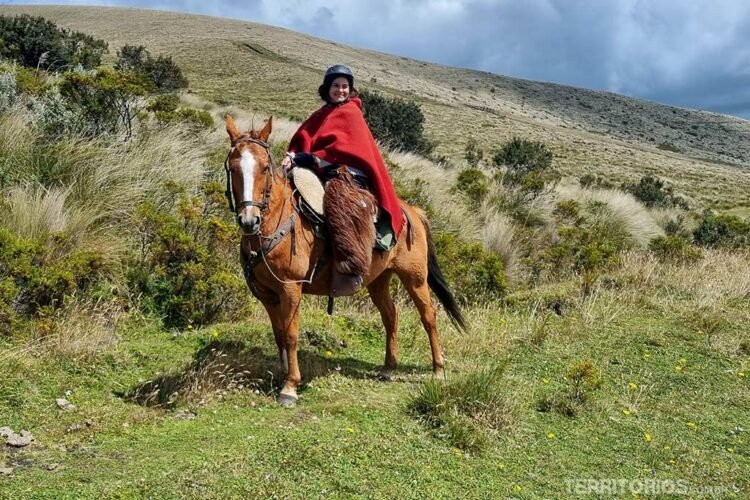 Mulher vestida com roupas para frio extremo com poncho vermelho e calça de pelos, monta cavalo no campo em frente ao Vulcão Cotopaxi