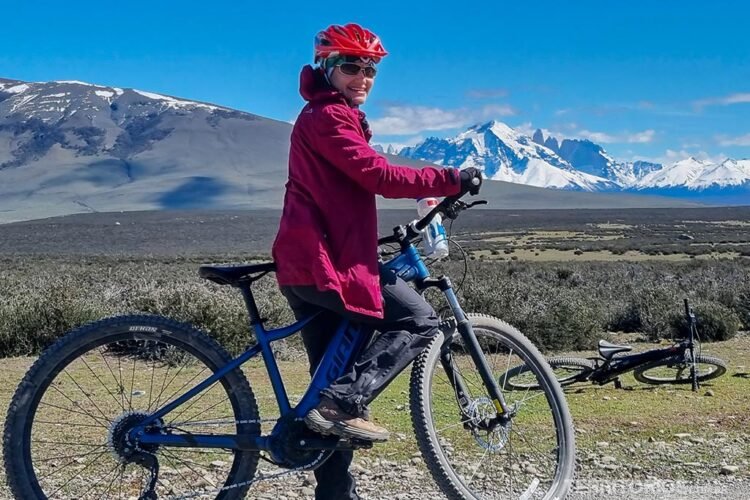 Mulher em cima de uma bicicleta veste jaqueta rosa com capuz, capacete de bicicleta, óculos e bandana. Ao fundo montanhas nevadas da Patagônia chilena