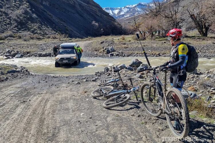 Mulher segura bicicleta e outras estão deitadas na estrada de terrada Patagônia chilena. Ao fundo, carro atravessa rio com pessoas penduradas.