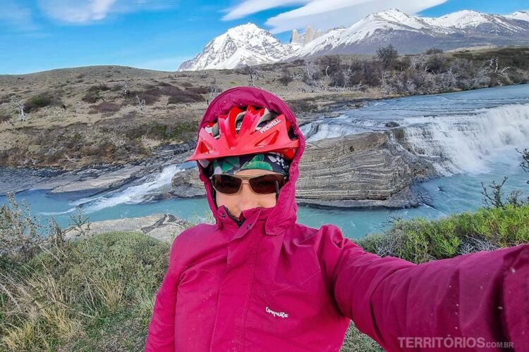 Mulher com frio veste jaqueta rosa com capuz, capacete de bicicleta, óculos e bandana. Ao fundo cachoeira de água do degelo vindo das montanhas da Patagônia chilena