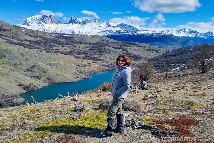 Mulher veste roupa cinza e protetores de tornozelo. Cabelos castanhos e óculos escuro.  Ao fundo lagoa azul, vegetação rasteira e as montanhas nevadas do Parque Nacional Torres del Paine
