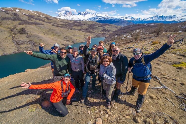 Grupo de pessoas celebra a trilha. Ao fundo montanhas nevadas, lago e ícone das Patagônia chilena: as Torres 