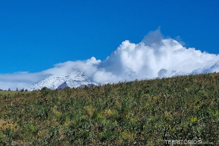 Vulcão Cotopaxi ao longe coberto por nuvens, campo verde e céu azul