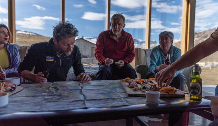 Pessoas sentadas ao redor da mesa comendo aperitivos. Ao fundo janelas com vista das montanhas e céu azul da Patagônia chilena