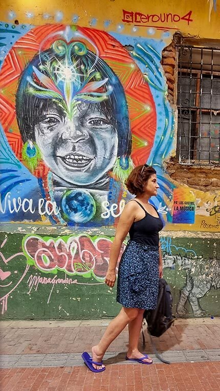 Mulher caminha em Santa Marta em frente a parede amarela com janela e grafite de indígena